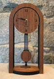 This shows a pendulum clock with a round top  above two straight legs of the clock, with a pendulum swinging in the middle. The motif is Walnut against black. The style is a solid-modern look with a mission-style pendulum and sides. This is the smaller size.