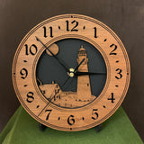 Round walnut clock with a lighthouse, moon and lightkeeper's house lasered in the face against a black background - 8" on easel