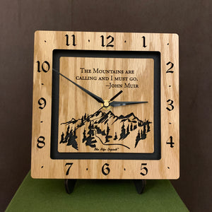 A square oak clock with the numbers on the outer square section, while on the inner square section a mountain and the words, "The Mountains are calling and I must go. -John Muir" are lasered in the wood. The concentric wood squares have a gap between them and are against a black background. 8" Size